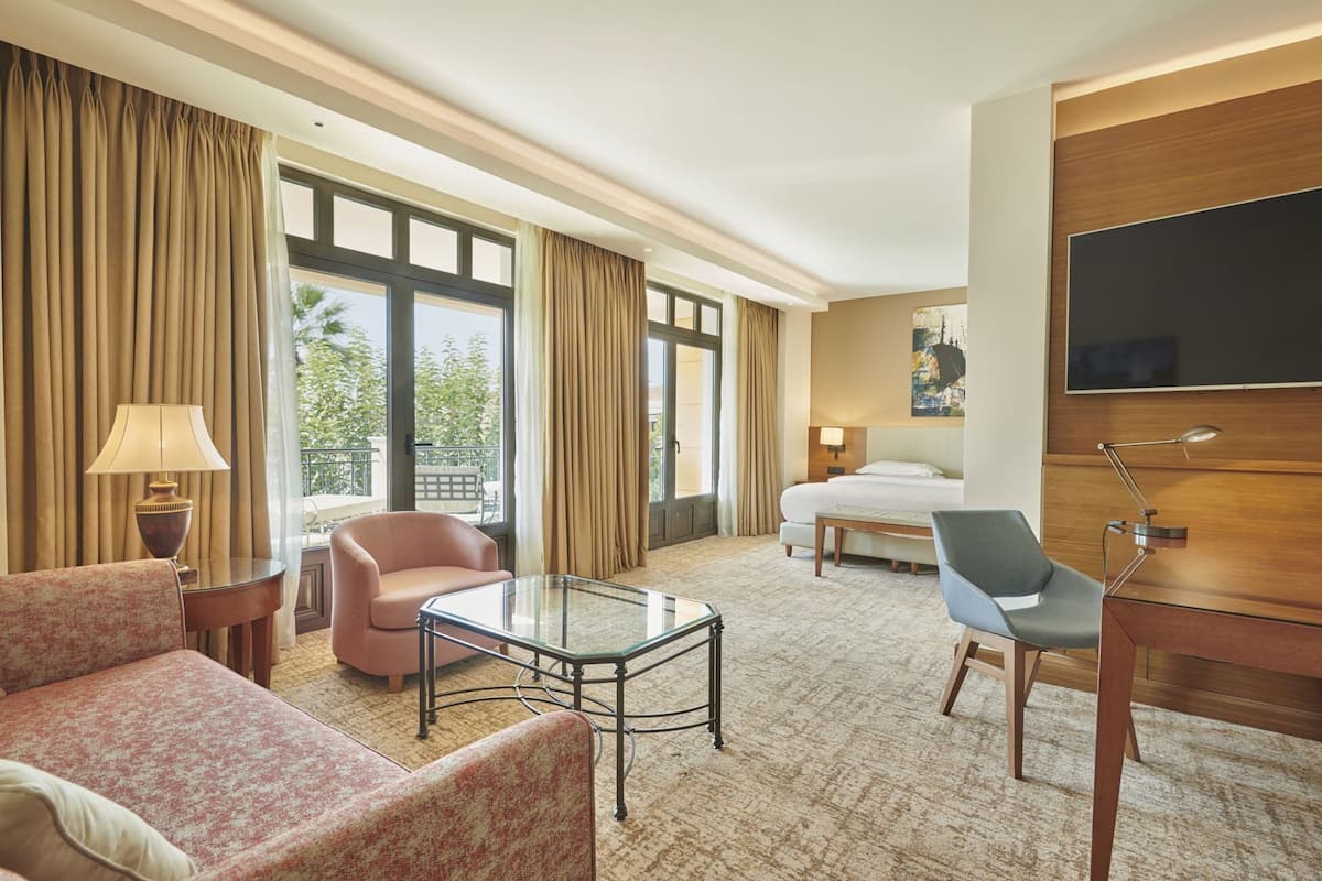 A hotel room with a bed, two chairs, a small table, a TV, and a balcony with curtains.