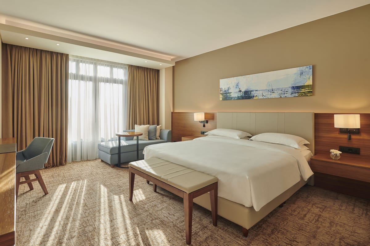 A hotel room with a large bed, a desk, a chair, a small table, and a painting on the wall.