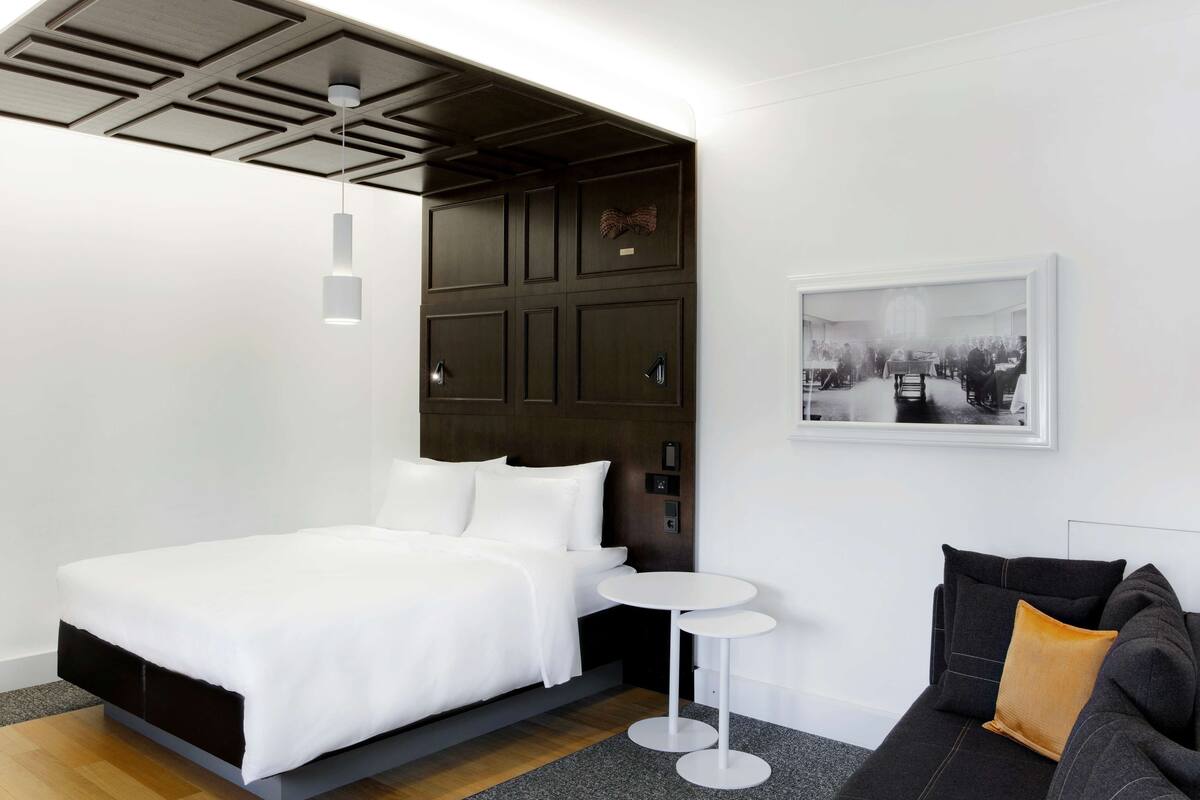 A modern hotel room with a large bed, a small round table, a sofa with cushions, a wooden headboard, and a framed picture on the wall.