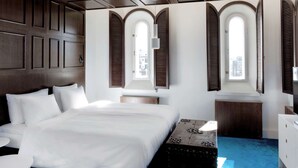 Habitación de hotel con una cama grande, cabecero de madera, una mesita redonda pequeña y un escritorio con lámpara. Hay tres ventanas con contraventanas, y la habitación tiene un diseño moderno.