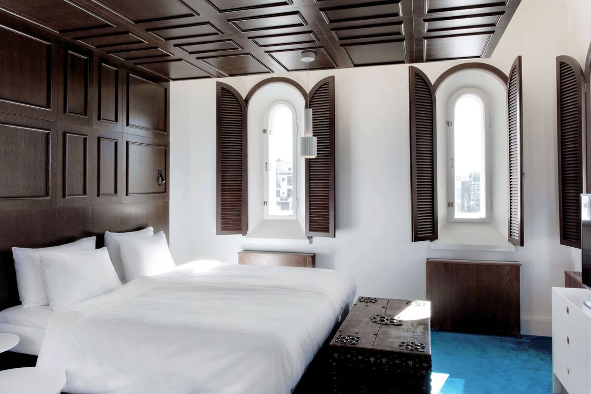 A hotel room with a large bed, a wooden headboard, a small round table, and a desk with a lamp. There are three windows with shutters, and the room has a modern design.