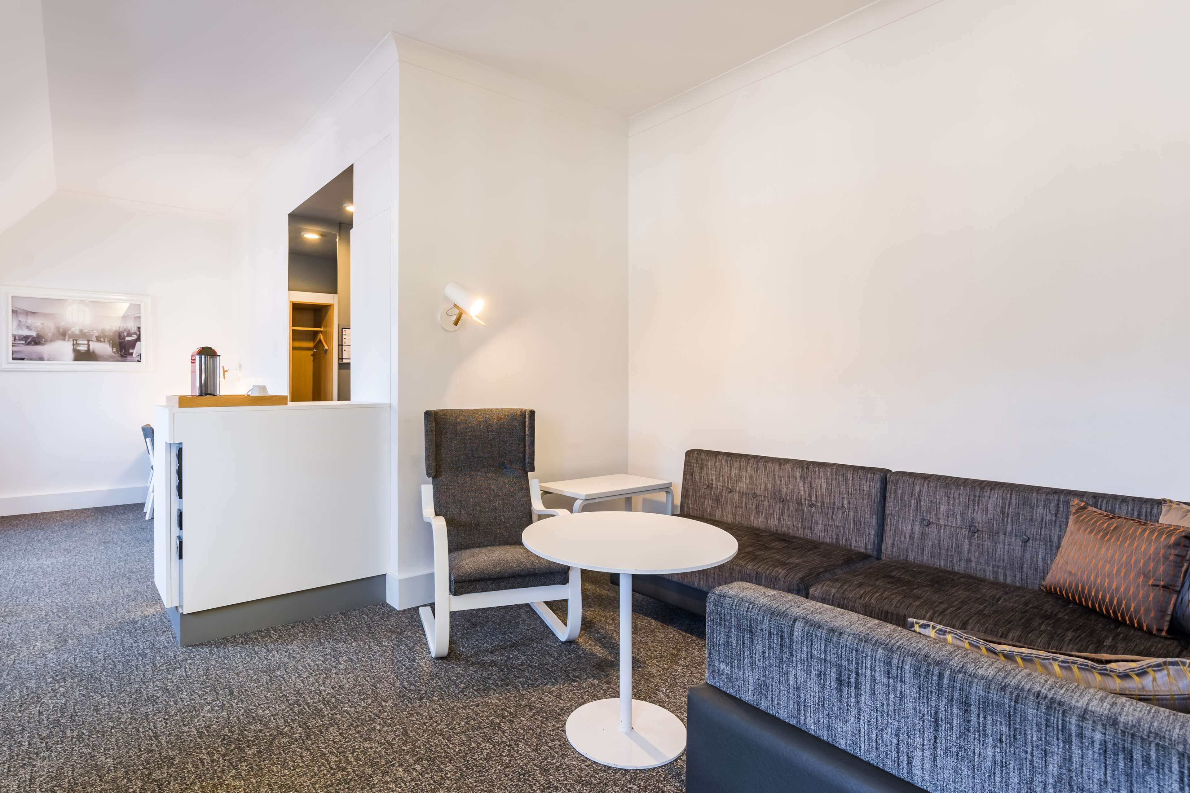 A modern hotel room with a grey sofa, a small round table, a grey armchair, and a white cabinet.
