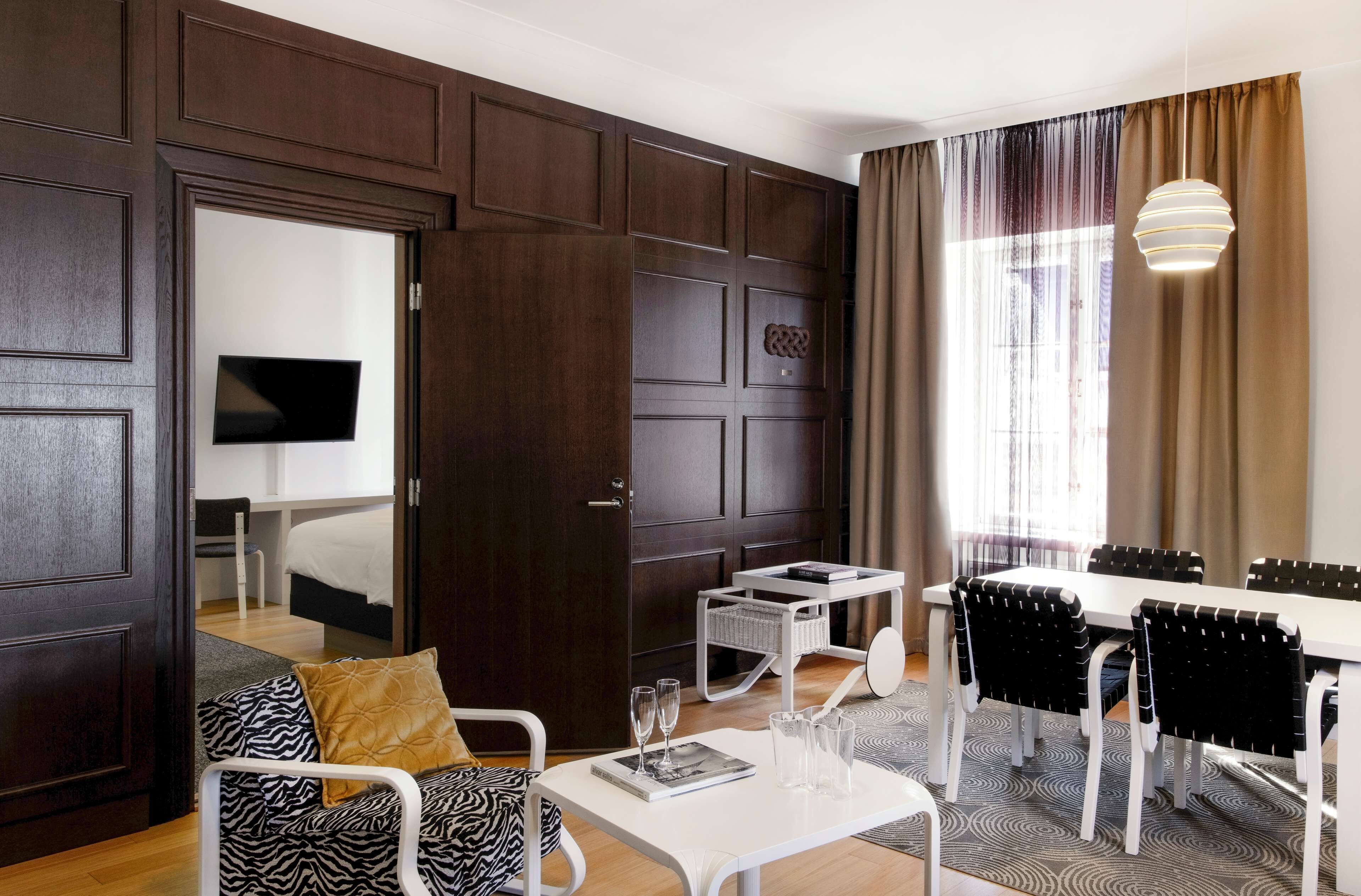 A modern hotel room with a dark wood panel wall, a white bed, a sofa with patterned cushions, a dining table with chairs, and a large window with curtains.