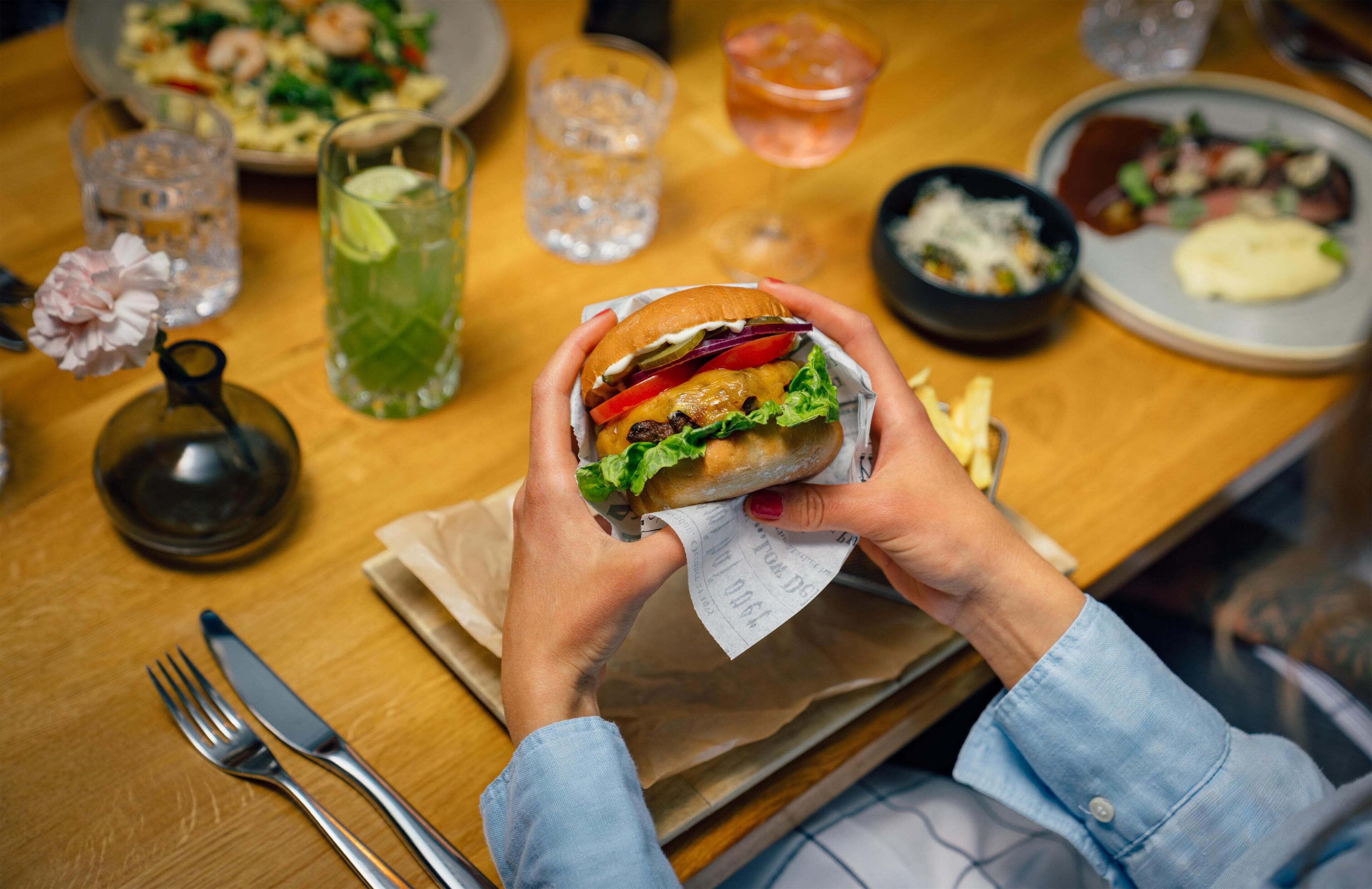 A person holding a sandwich with a paper wrapper, a glass of iced drink, and a plate of food on a wooden table.