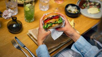 A person holding a sandwich with a paper wrapper, a glass of iced drink, and a plate of food on a wooden table.