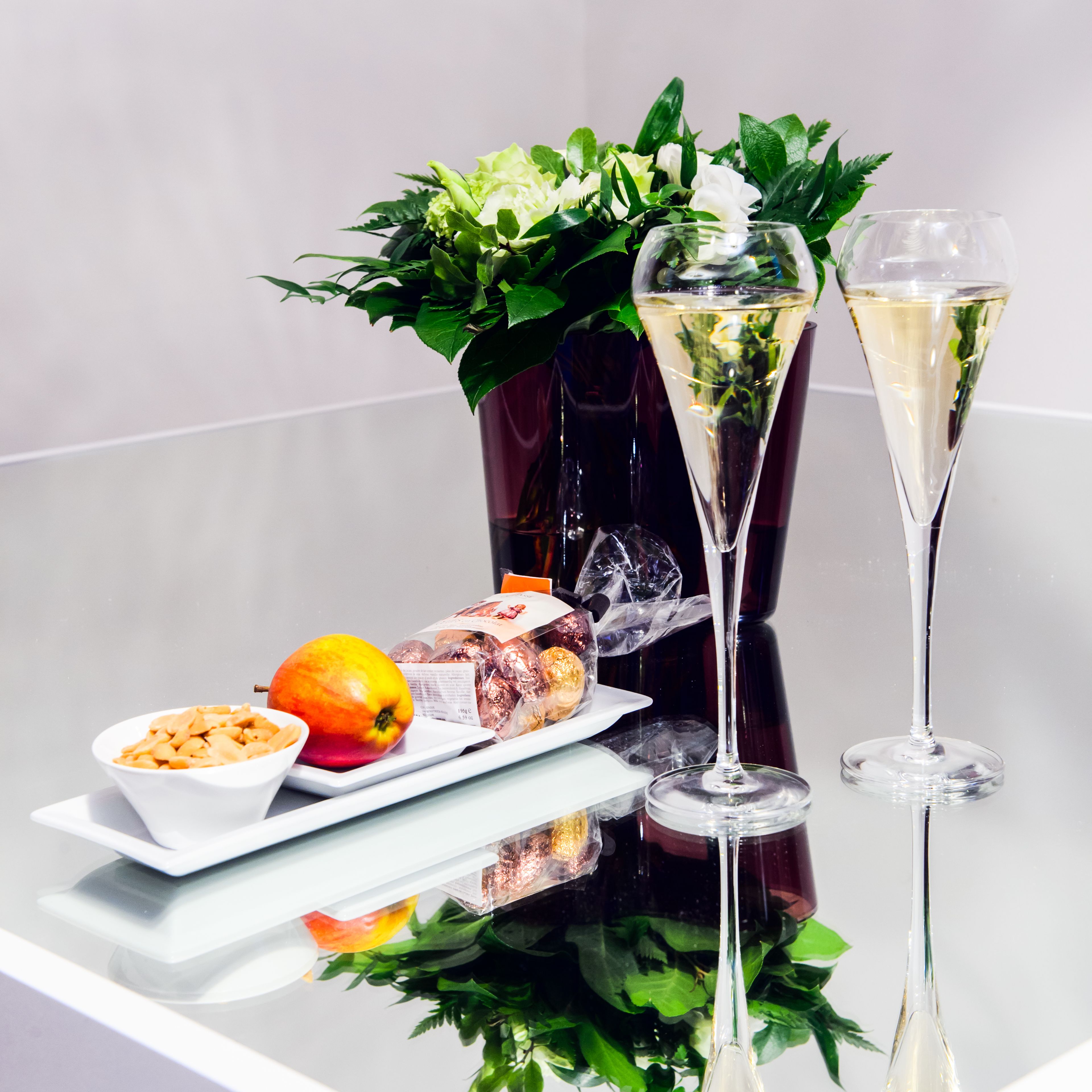 Un tavolo con un bicchiere di champagne, un bicchiere d'acqua, una ciotola di snack, un frutto e un bouquet di fiori.
