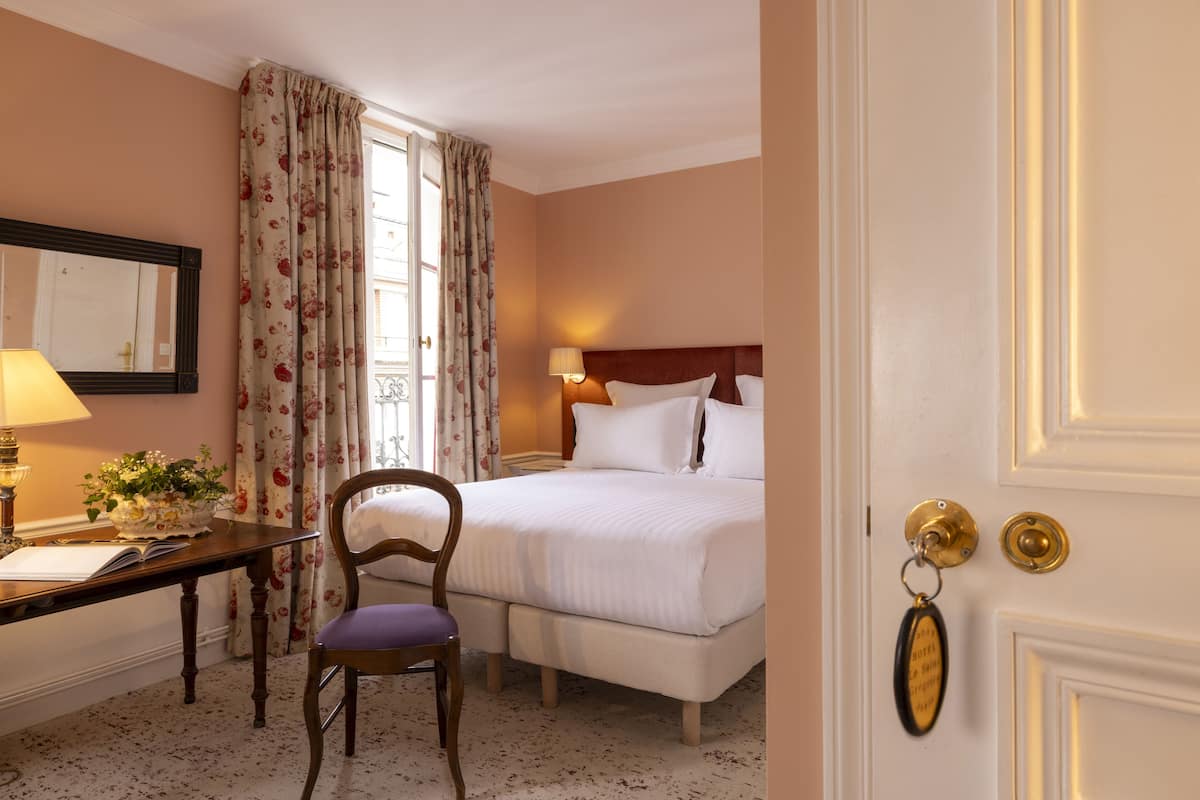 A hotel room with a bed, a desk with a lamp, a chair, and a window with curtains.