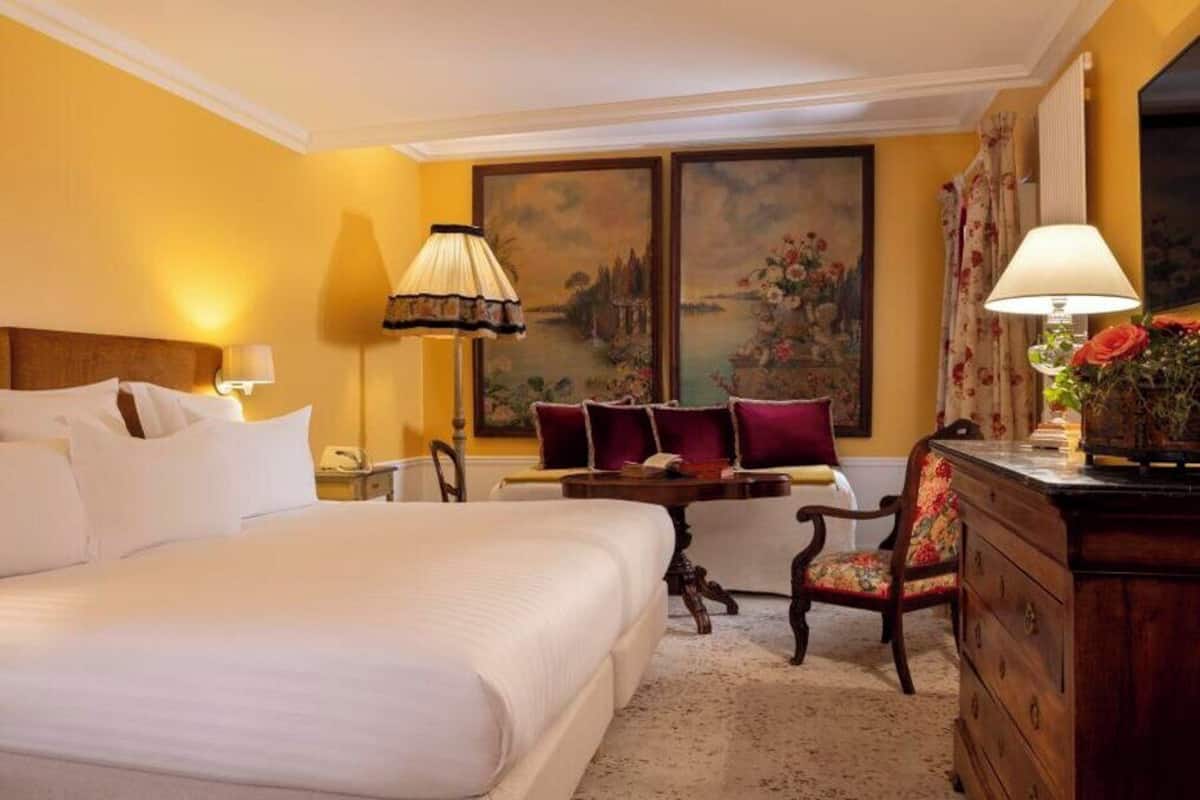 A hotel room with a large bed, a desk, a chair, and a dresser. There are two paintings on the wall and a lamp on the desk.