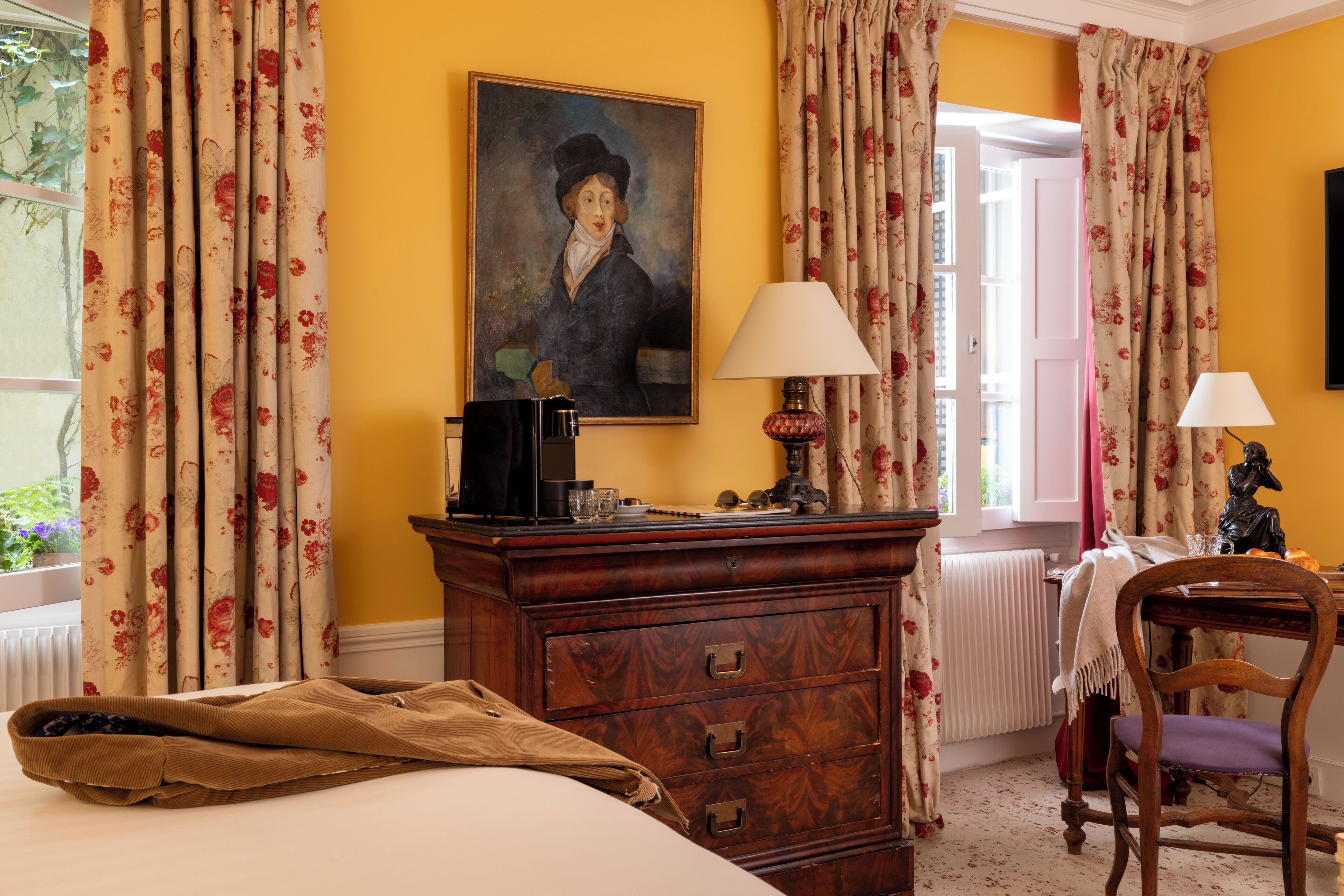 A hotel room with a bed, a wooden dresser, a desk with a lamp, a chair, and a painting on the wall.