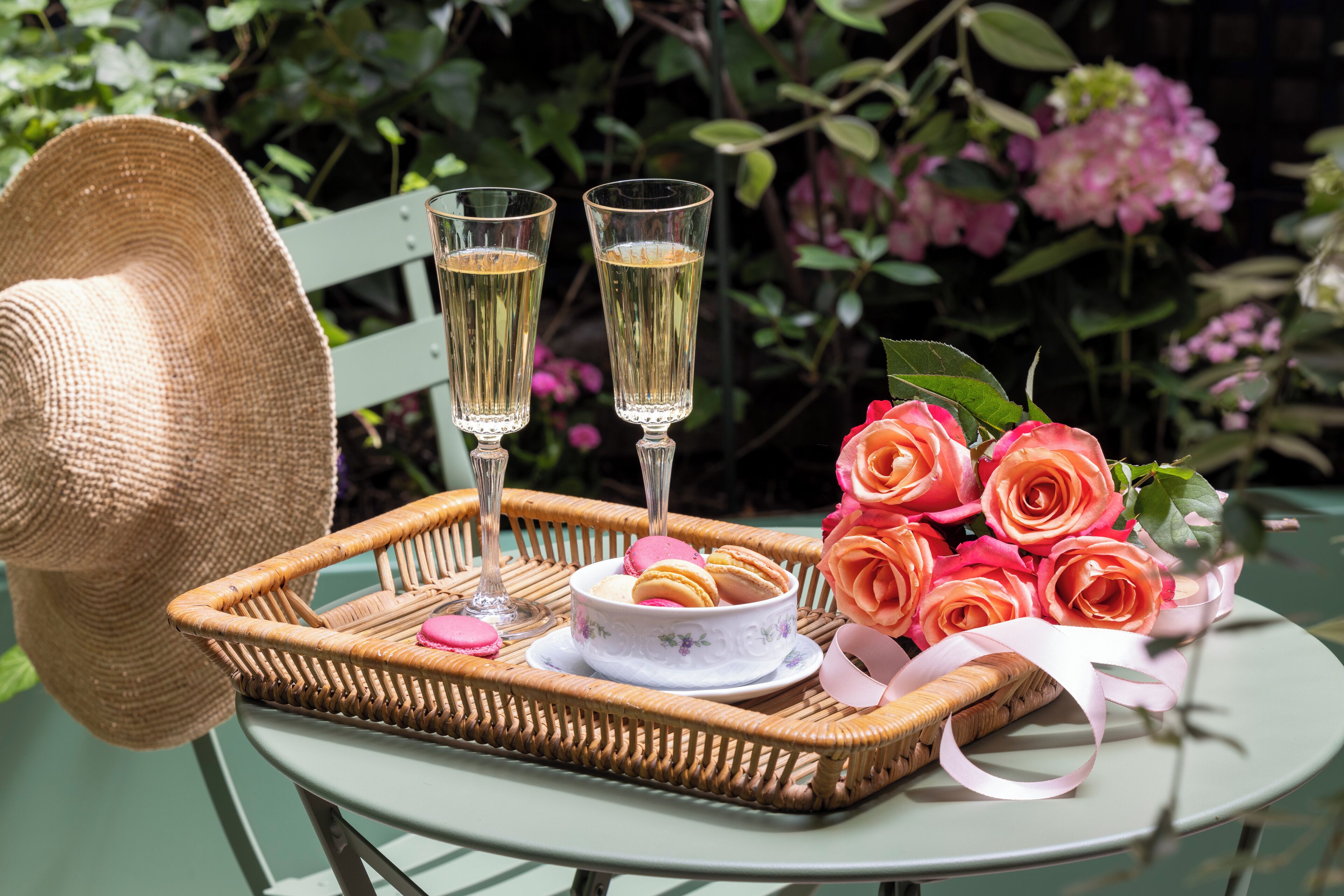 A wicker tray with two glasses of a clear liquid, a bowl of macarons, and a bouquet of pink roses on a table.