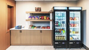 A kitchenette with two black refrigerators stocked with beverages and snacks, a wooden cabinet with various food items, and a shelf with packaged goods.