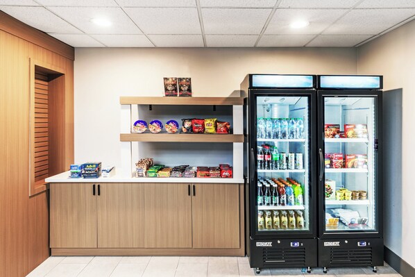 A kitchenette with two black refrigerators stocked with beverages and snacks, a wooden cabinet with various food items, and a shelf with packaged goods. - Holiday Inn Express Breaux Bridge by IHG (Breaux Bridge)