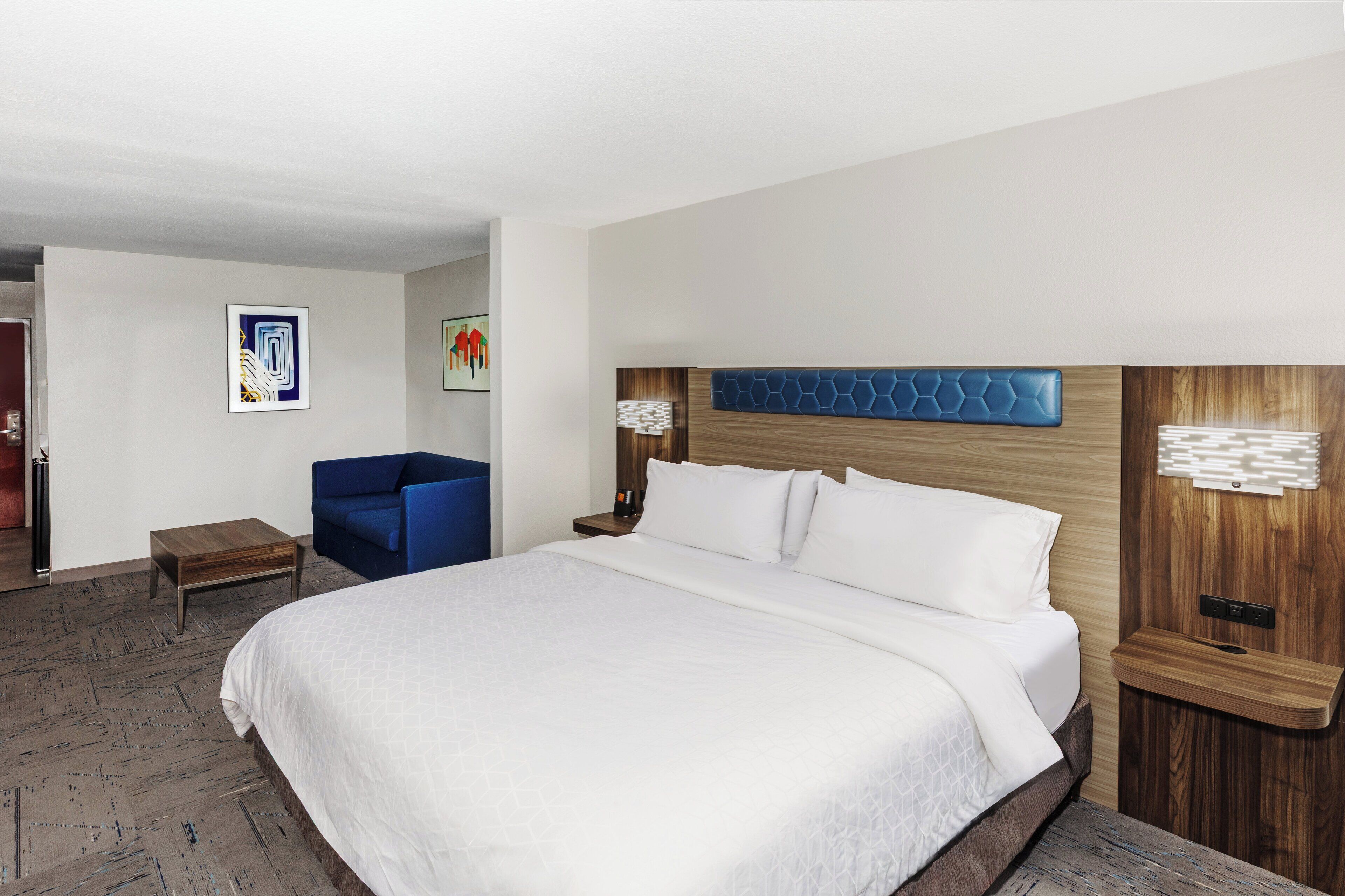 A modern hotel room with a large bed, a blue armchair, and a wooden headboard.
