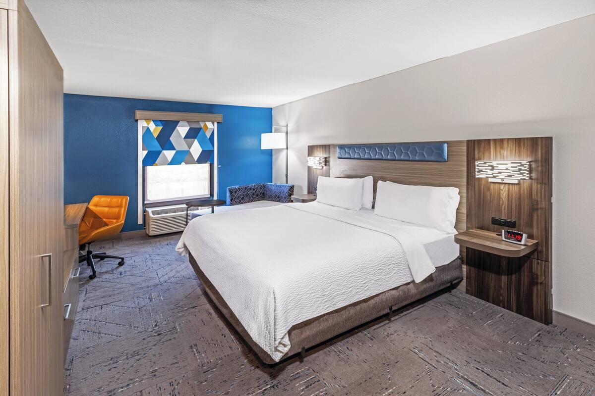 A hotel room with a large bed, a desk, an orange chair, and a window with a geometric curtain.