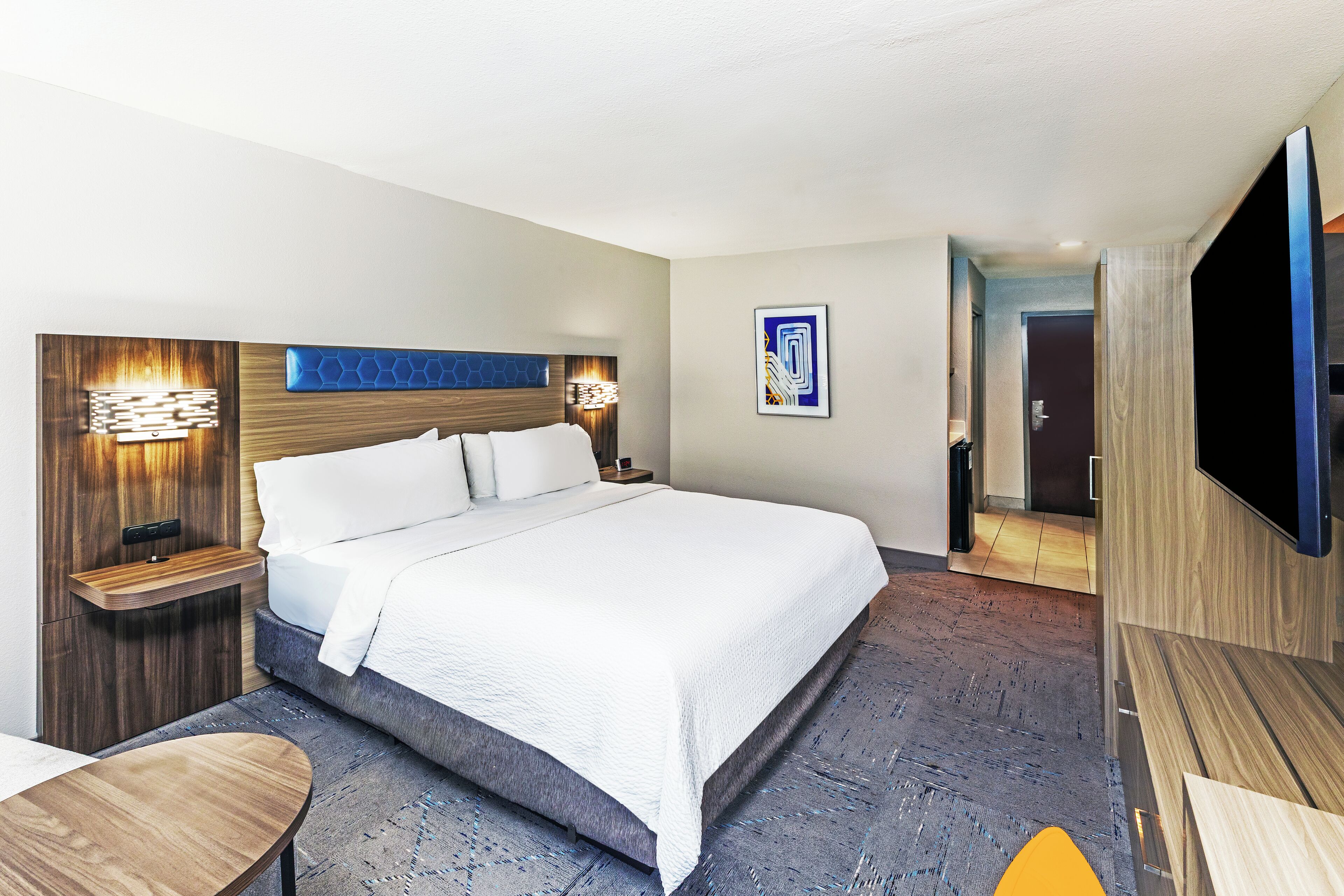 Una habitación de hotel moderna con una cama grande, una mesita de noche de madera, un televisor de pantalla plana y una obra de arte colgada en la pared.