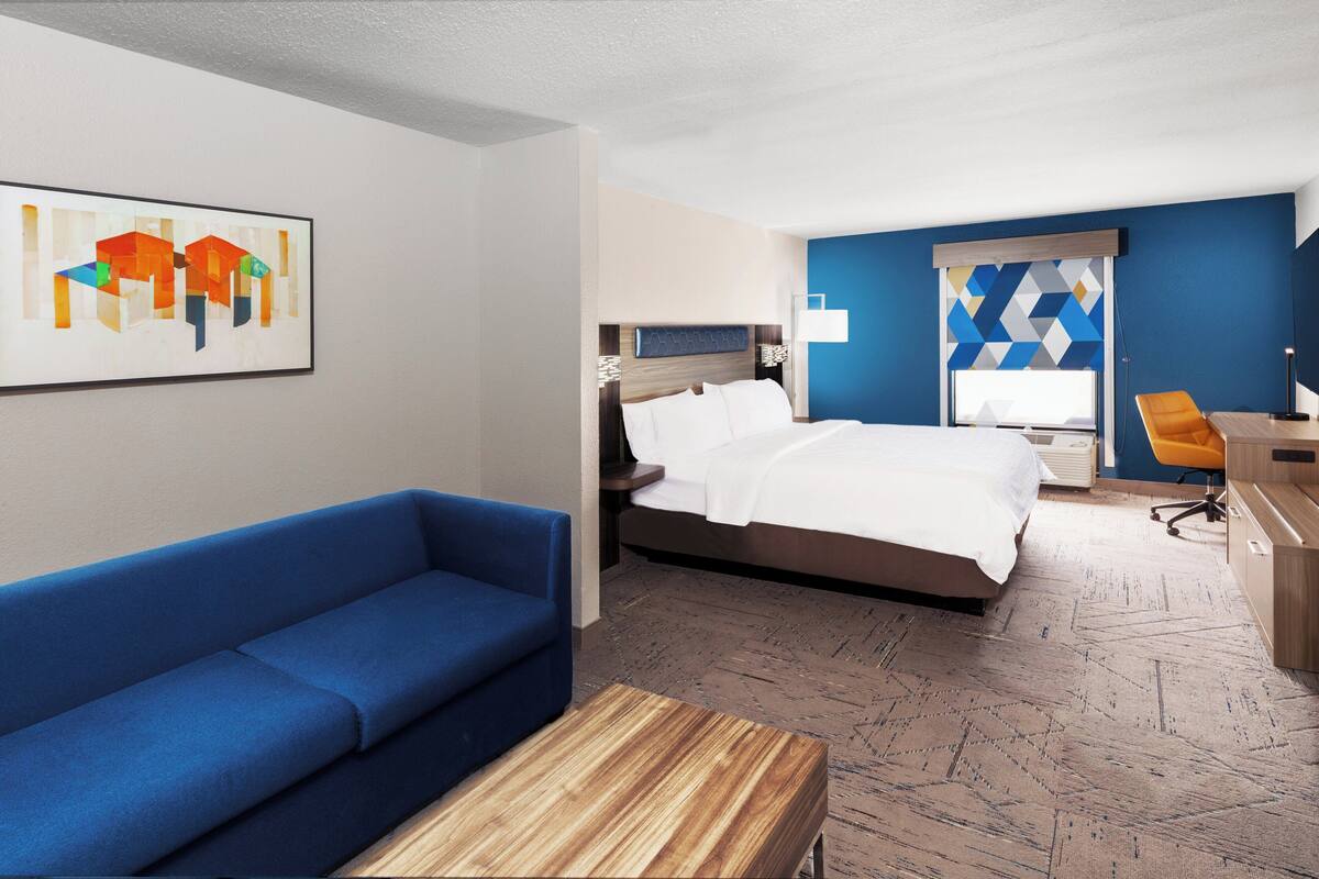 A modern hotel room with a bed, a blue sofa, a wooden coffee table, and a desk with a chair.