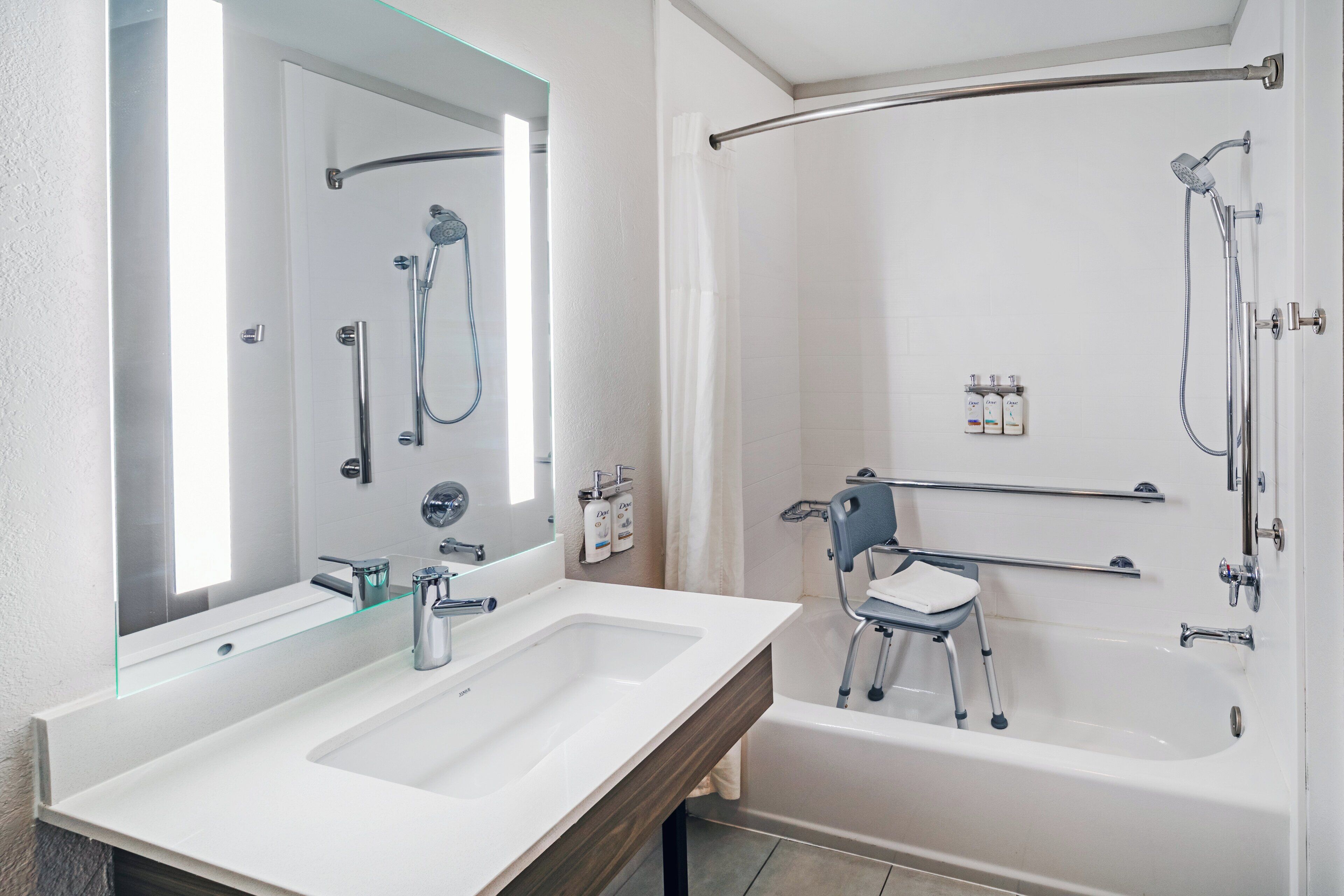 A modern bathroom with two sinks, a large mirror, a shower with a grab bar, and a wheelchair-accessible bench.