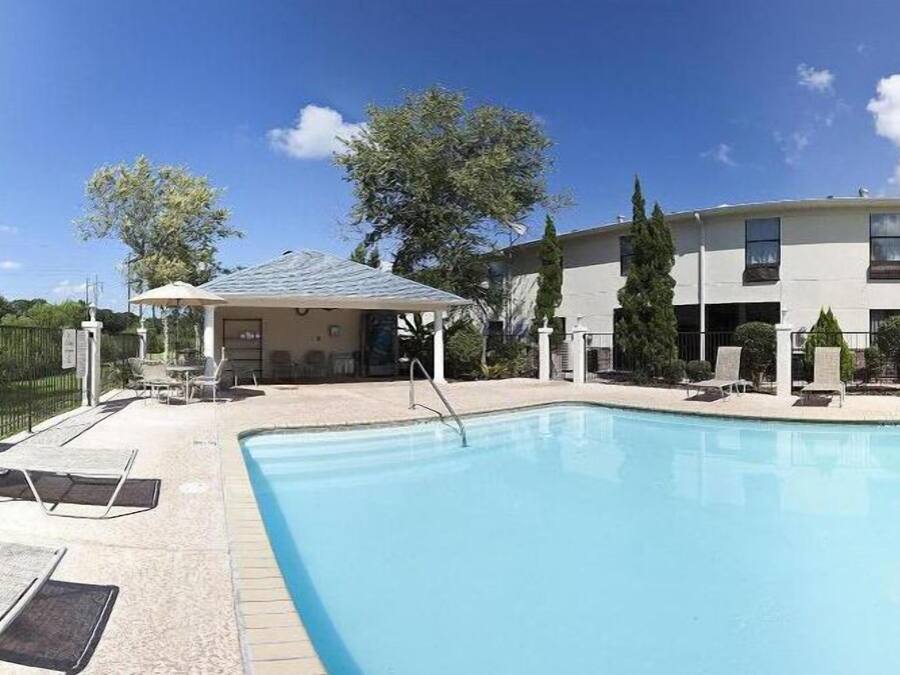 A swimming pool with lounge chairs and a covered seating area, surrounded by a building with multiple windows and a fenced garden.