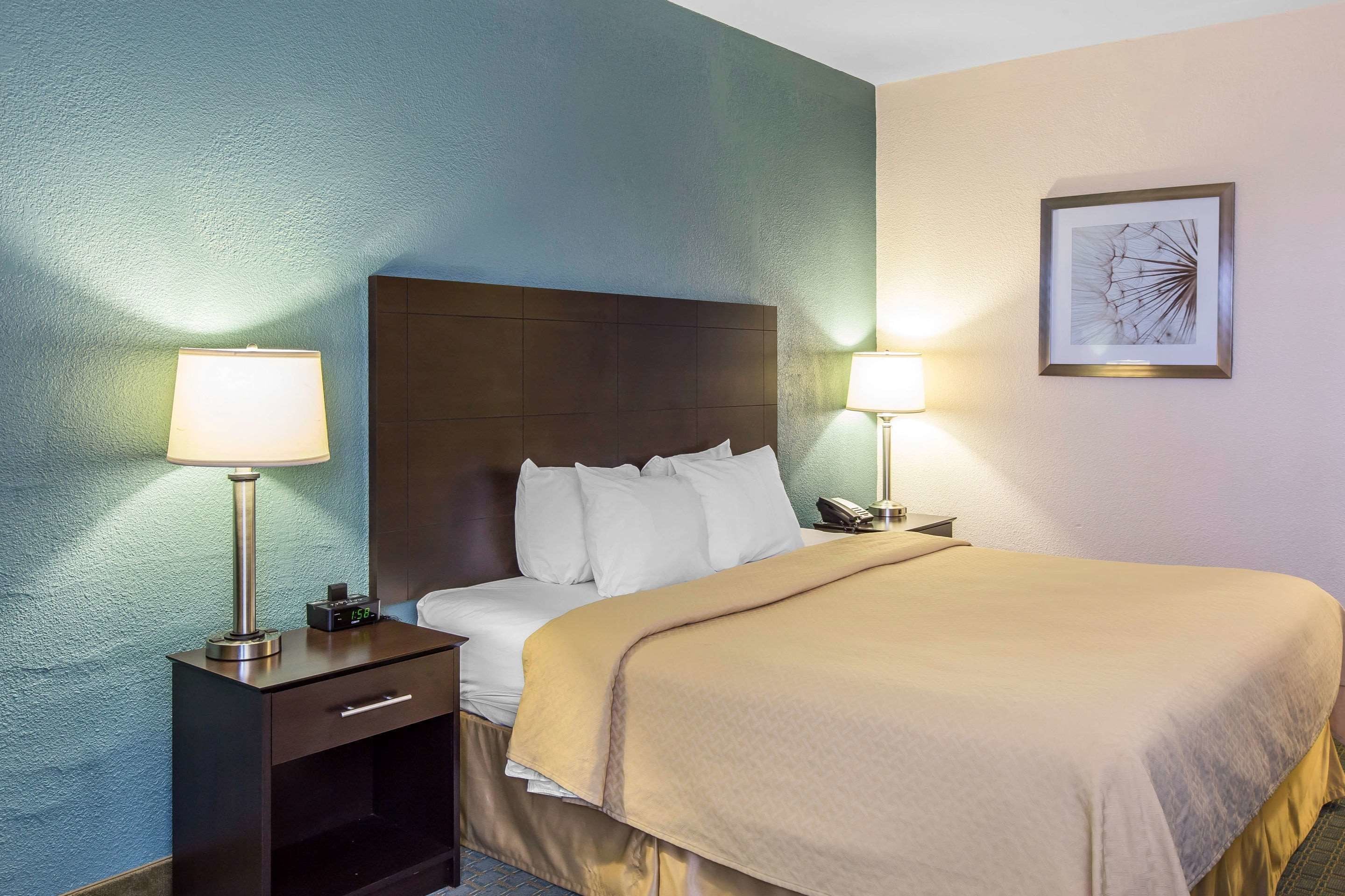 Quarto de hotel com uma cama, abajures, mesa de cabeceira e uma obra de arte emoldurada na parede.