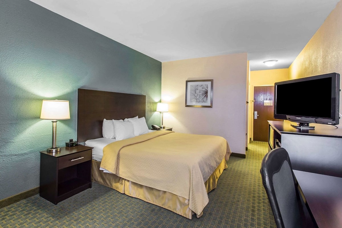 Quarto de hotel com cama, luminárias de cabeceira, escrivaninha com abajur, televisão e um quadro na parede.