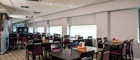 A conference room with multiple tables and chairs, a large screen, and a kitchen area in the background.