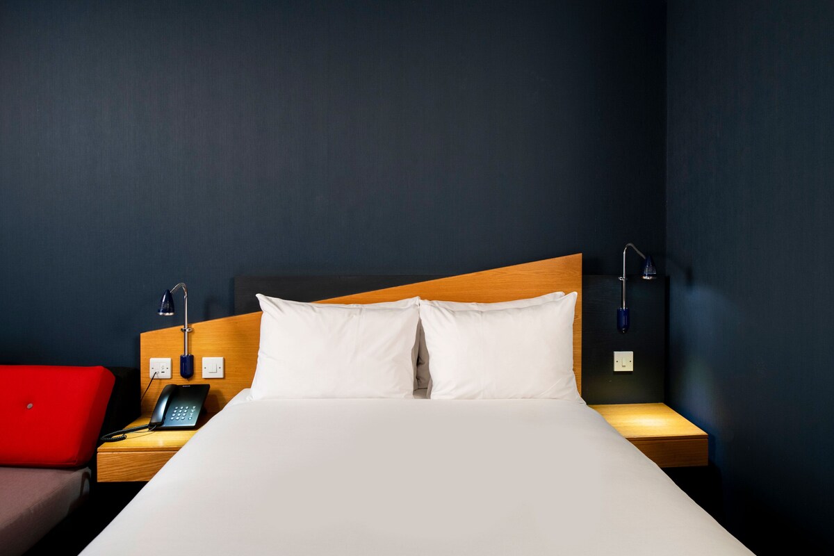 A hotel room with a bed, a nightstand, a red chair, and a phone.