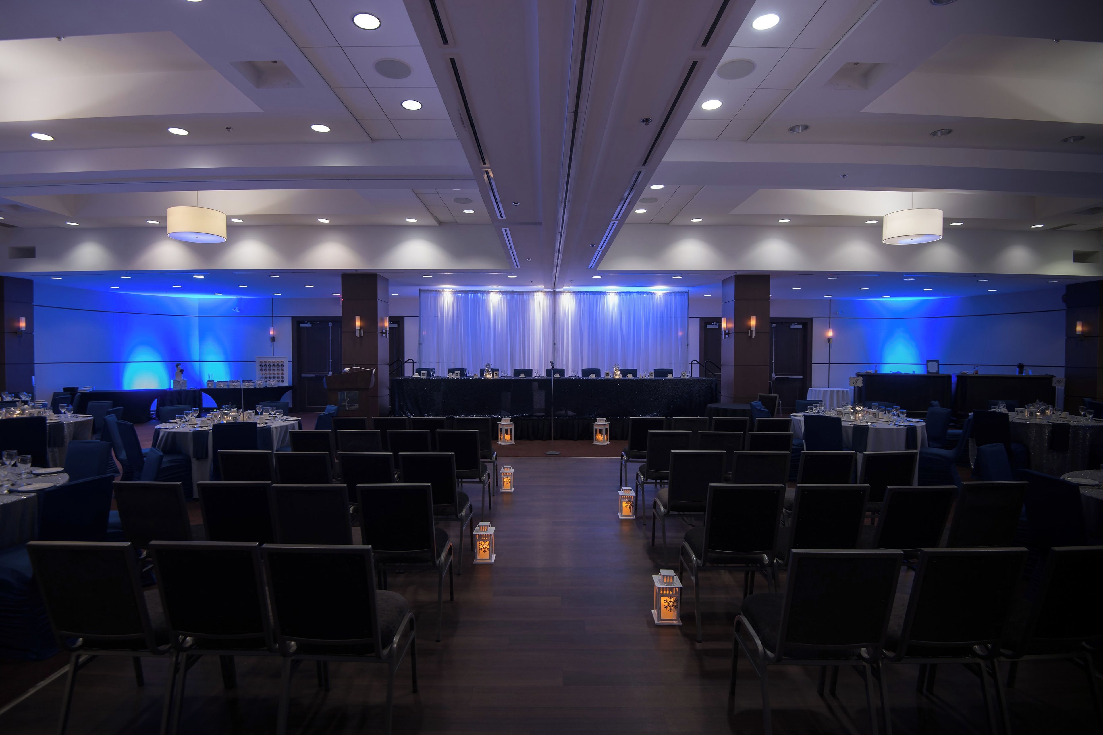 A conference room with rows of tables and chairs, a stage with a blue curtain backdrop, and recessed ceiling lighting.