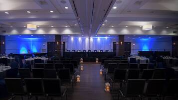 A conference room with rows of tables and chairs, a stage with a blue backdrop, and recessed ceiling lighting.