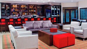 Un espace bar moderne avec un coin salon comprenant un canapé et des fauteuils, un comptoir de bar où sont exposées diverses bouteilles, et un élément décoratif rouge sur le sol.