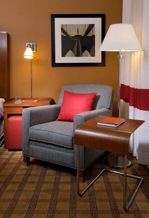 Una habitación de hotel con un sillón gris, un cojín rojo, una mesita de madera con un libro, una lámpara de pie y un cuadro en la pared.