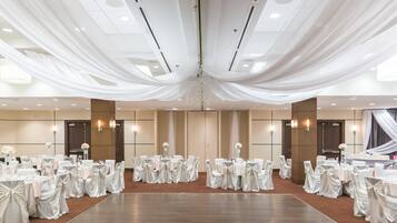 A large, elegantly decorated banquet hall with rows of round tables, each set with white tablecloths and chairs, and a central chandelier.