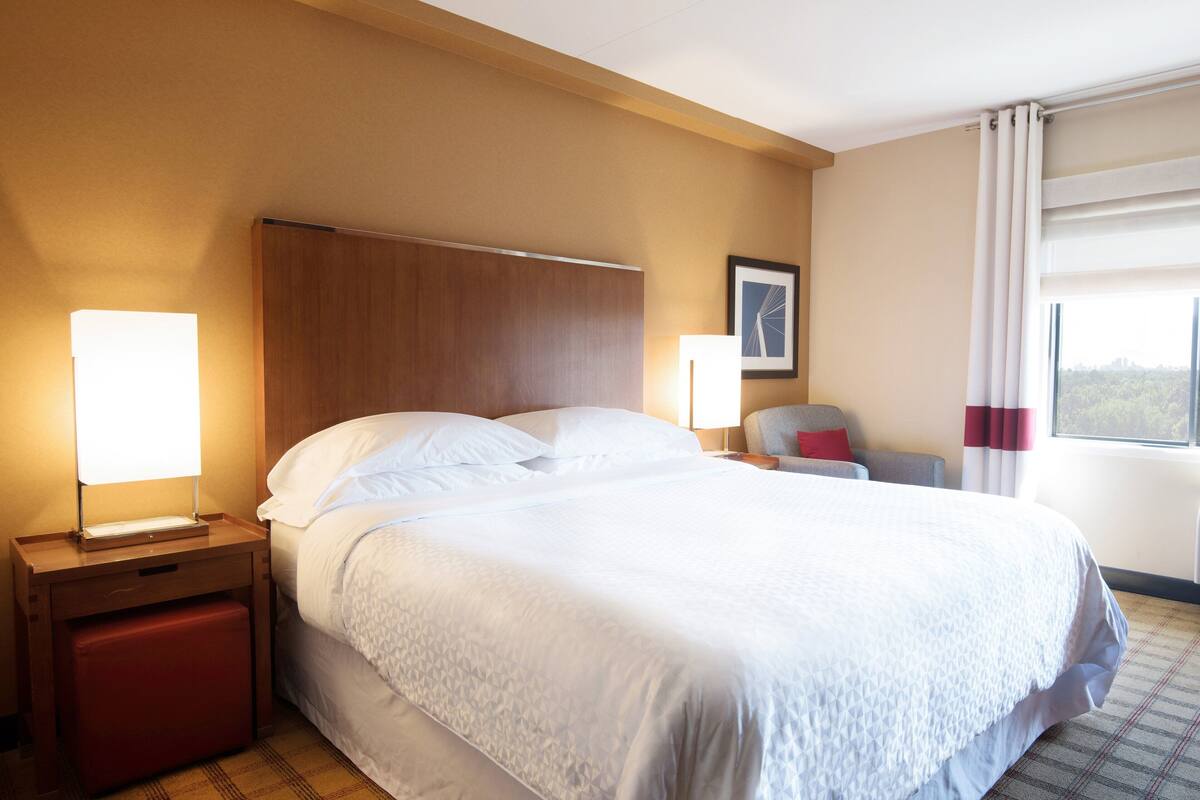 Quarto de hotel com uma cama grande, duas luminárias de cabeceira, uma mesa de cabeceira, uma cadeira, uma janela com cortinas e um quadro na parede.