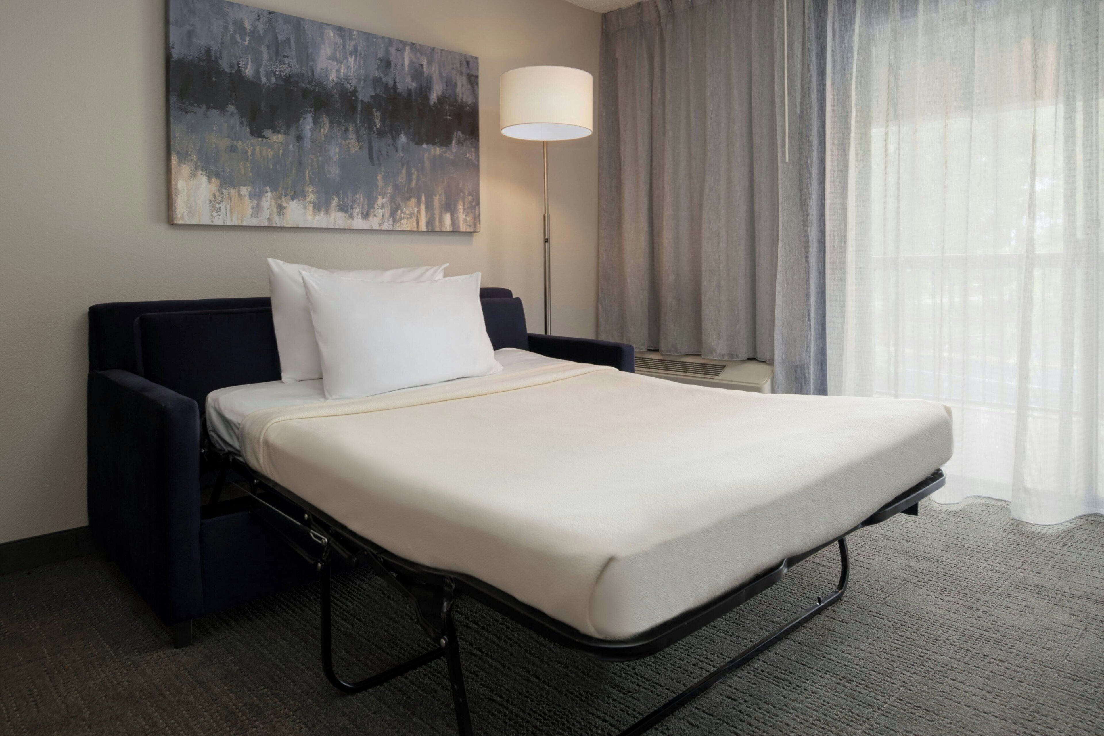 A single bed with a white mattress and a headboard, a bedside table with a lamp, and a painting on the wall.
