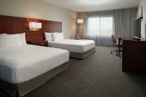 A hotel room with two beds, a desk, a chair, a TV, and a lamp.