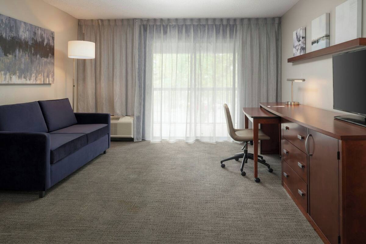 Une chambre d’hôtel avec un canapé bleu foncé, un bureau avec des tiroirs, une chaise, une grande fenêtre avec des rideaux légers, une lampe sur pied et un téléviseur à écran plat.