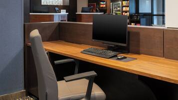 A modern office space with a wooden desk, a computer, a chair, and a reception area with a refrigerator and snacks.