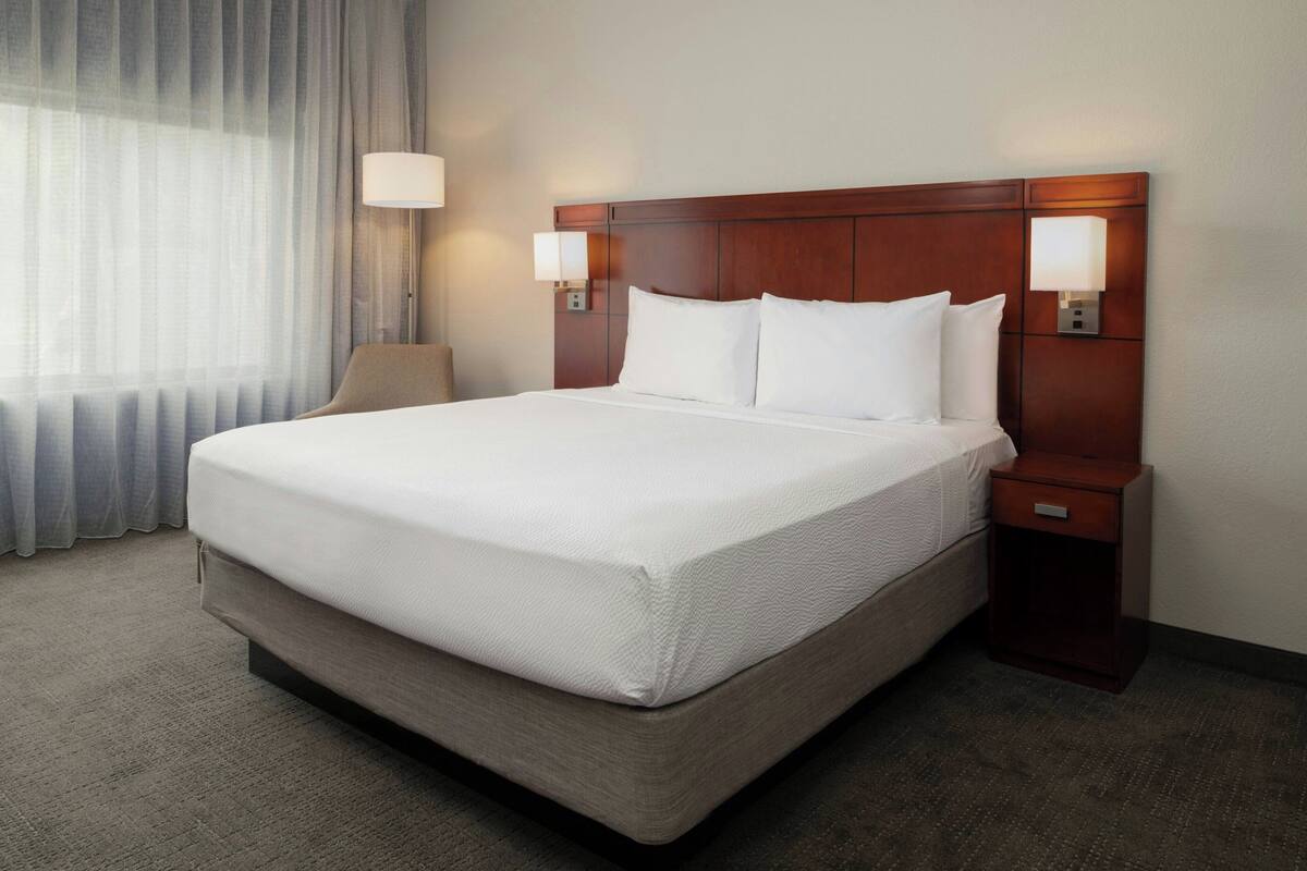 A hotel room with a large bed, two bedside tables with lamps, a nightstand, a chair, and a window with curtains.