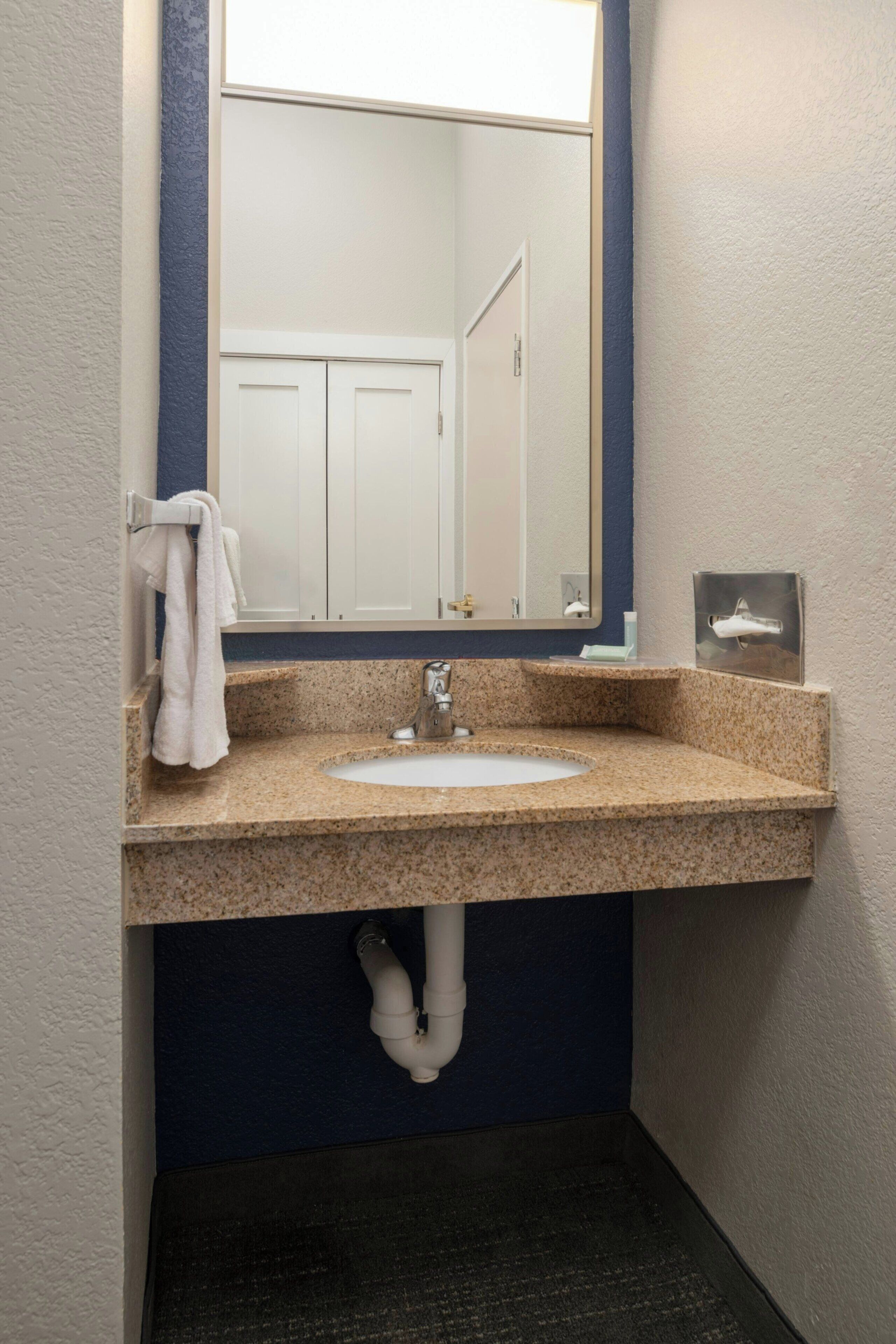 Une salle de bain avec un lavabo doté d’un plan de travail en granit, un miroir, un porte-serviettes et une porte visible au fond.