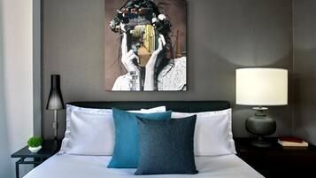 Une chambre d’hôtel avec un lit, des oreillers, une table de chevet, une lampe, un tableau encadré et une petite plante.