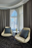 Ein Hotelzimmer mit zwei Ledersesseln, einem kleinen runden Tisch mit einer Flasche und Gläsern sowie einem großen Fenster mit Vorhängen.