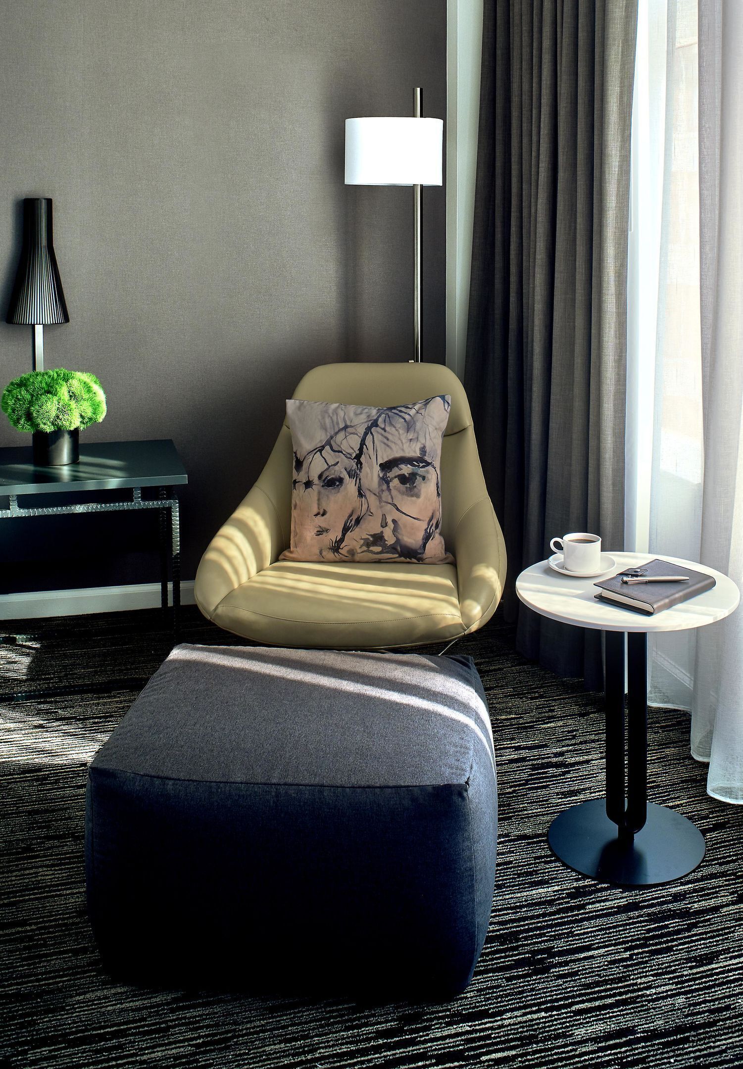 A modern hotel room with a sofa, ottoman, lamp, table with a cup and notebook, and a plant on a side table.