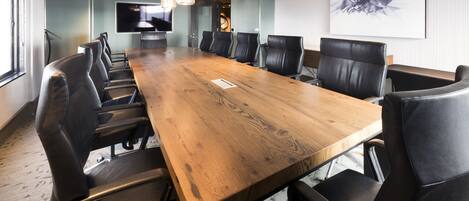 A conference room with a long wooden table, black leather chairs, and a large flat-screen TV on the wall.