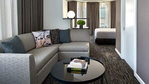 Habitació d'hotel moderna amb un sofà seccional gris, una taula de centre rodona amb llibres, una finestra gran amb cortines i un llit al fons.
