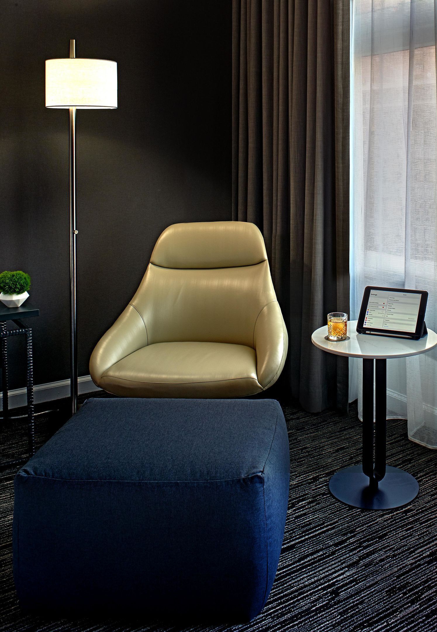 A hotel room with a leather armchair, a small table with a tablet, a lamp, and a view of the outdoors through sheer curtains.
