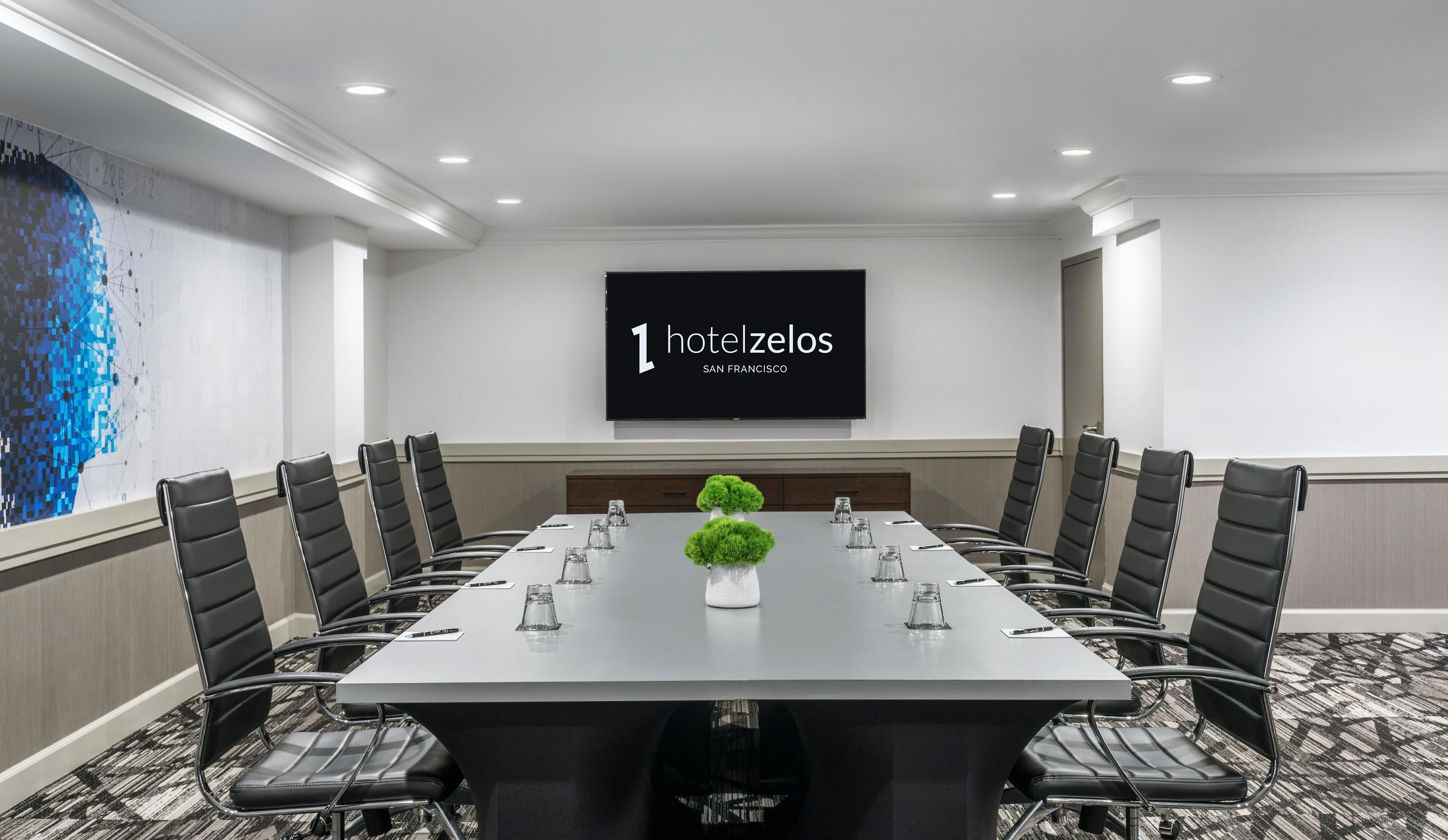 A conference room with a long table, black chairs, a projector screen, and a decorative plant.