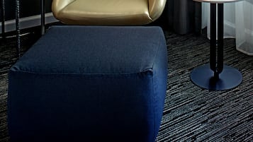 A modern hotel room with a leather armchair, a small round table with a tablet, a lamp, and a view of the outdoors through sheer curtains.