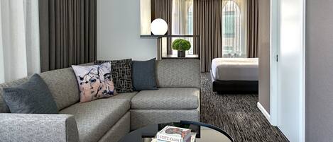 A modern hotel room with a grey sectional sofa, a round coffee table with books, a bed, a window with curtains, and a small potted plant.