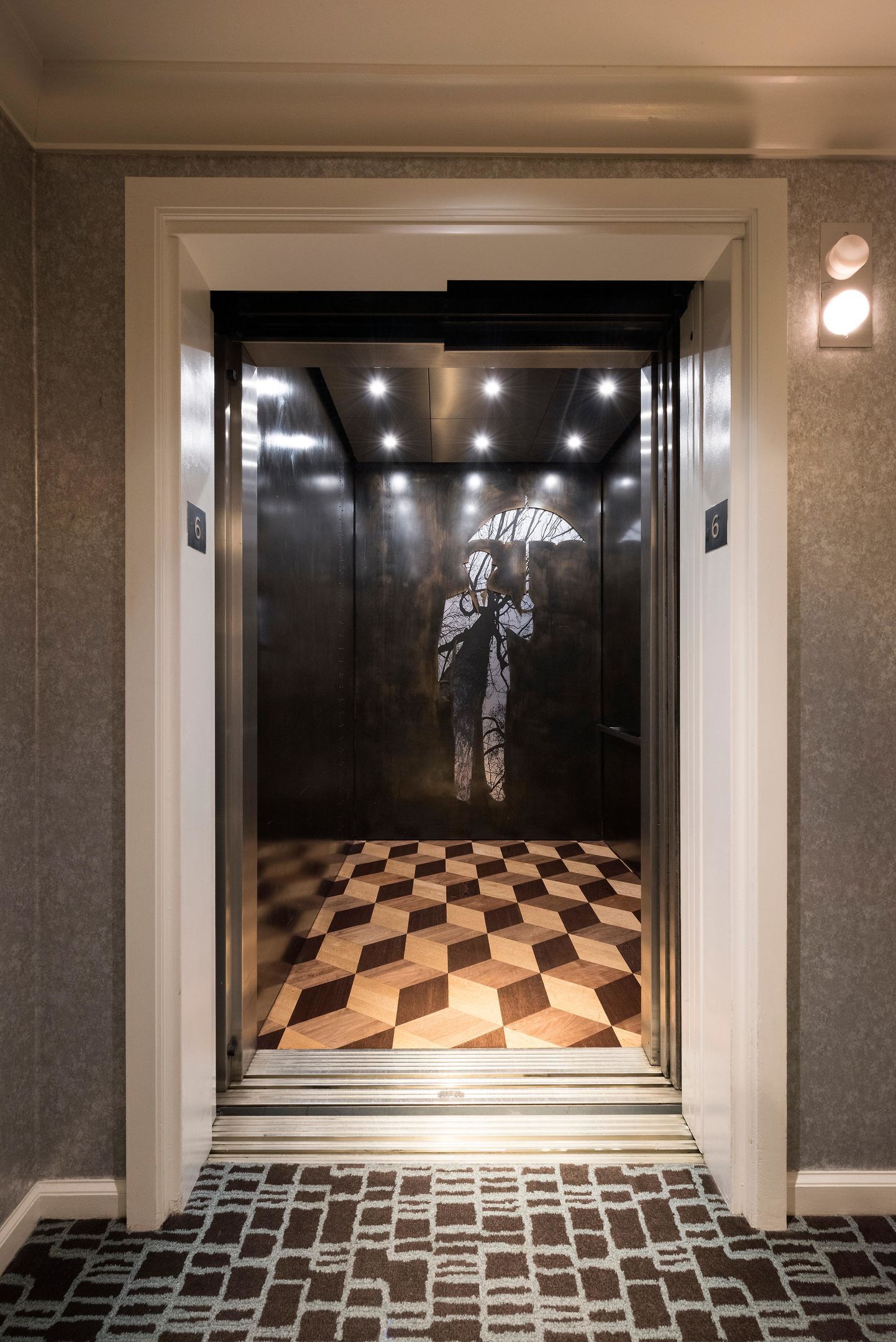 An elevator with a geometric patterned floor, a metallic interior, and a wall-mounted light fixture.