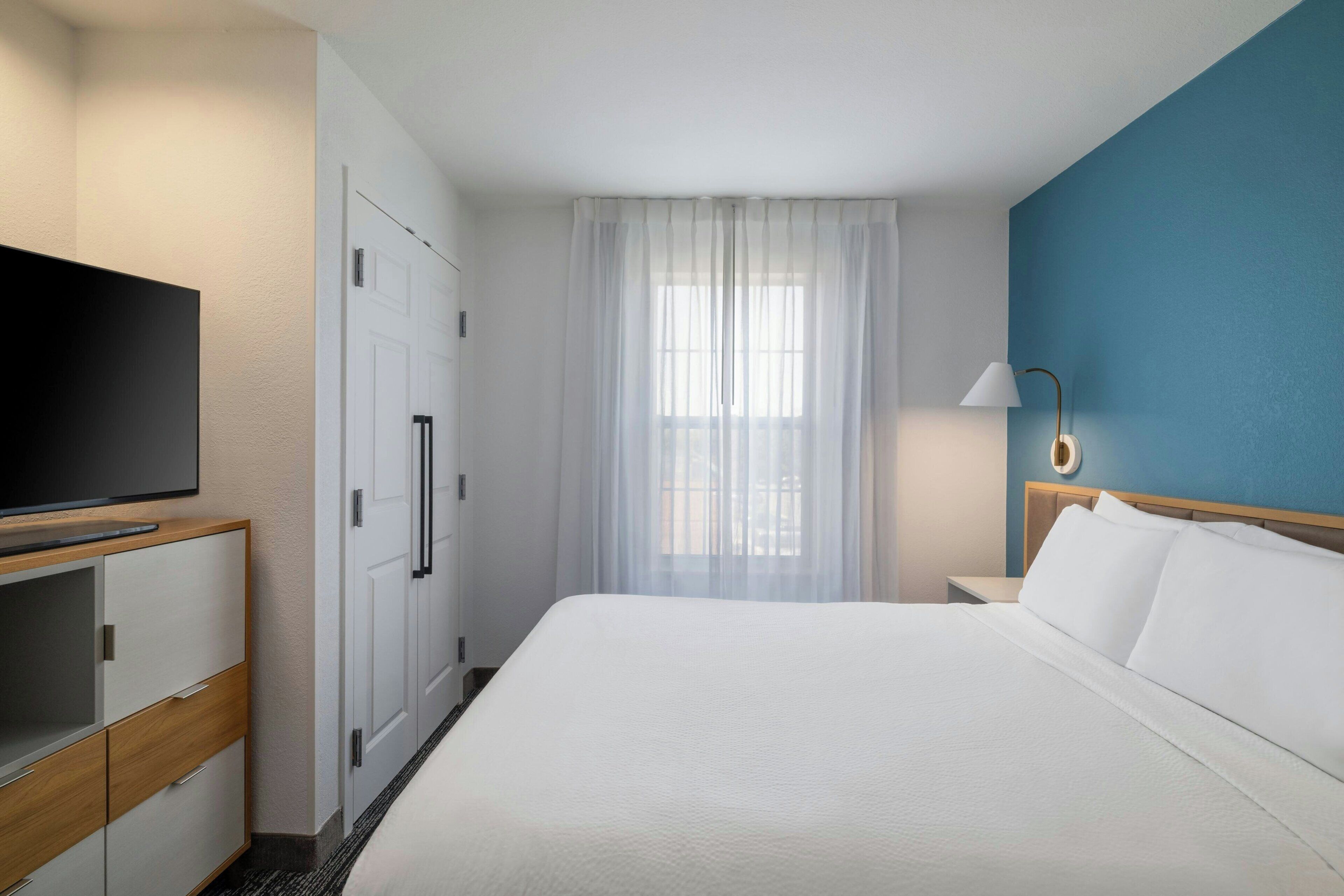 A hotel room with a bed, a TV, a wardrobe, a window with curtains, and a lamp.
