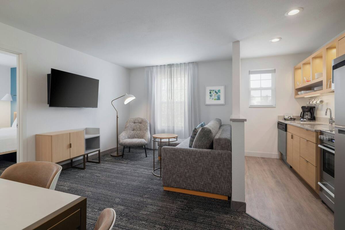 Et moderne hotelværelse med sofa, lænestol, spisebord, fladskærms-tv, tekøkken og et stort vindue med gardiner.